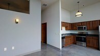This single-story space makes a strong architectural impression through its dramatically tall ceilings, which lend an open, airy quality to the interior. The polished concrete floors run continuously throughout, providing a sleek, industrial aesthetic that complements the clean white walls and crisp lines of the space.

The kitchen is positioned along one wall and is defined by warm medium-toned wood cabinetry that extends both above and below the dark countertops, offering generous storage. A stainless steel sink is centrally placed beneath upper cabinets, and stainless steel appliances, including a dishwasher and range, integrate seamlessly into the layout. A pendant light fixture above the kitchen area adds a functional and decorative touch.

A structural column and partial wall divide the kitchen from the adjacent open living area, creating a subtle sense of zone separation while preserving the spacious feel. A doorway visible from the main area leads to additional rooms beyond. High windows near the ceiling allow natural light to filter in from above, enhancing the vertical drama of the space. Overall, the design conveys a modern, utilitarian character with clean, unfussy finishes throughout.