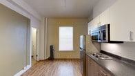 This single-floor unit offers a thoughtfully arranged living space that combines modern conveniences with classic architectural charm. The kitchen is a standout feature, equipped with stainless steel appliances including a refrigerator, built-in microwave, and a coil-burner cooktop, all complemented by dark granite-style countertops and contemporary two-tone cabinetry in cream and warm wood tones. Light hardwood flooring runs throughout, adding warmth and continuity to the space. A large window with white blinds allows natural light to fill the room, brightening the soft yellow-toned walls. A classic cast-iron radiator beneath the window adds a charming, vintage character consistent with older building construction. A structural column near the window is a notable architectural detail, hinting at the building's historic framework. A hallway visible from the kitchen leads to additional rooms, suggesting a practical and connected layout. The overall ambiance is clean, functional, and welcoming, with a blend of updated finishes and period details that give the space a distinctive character well-suited for everyday living.
