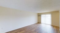 This single-floor space presents a clean, open layout with a focus on simplicity and functionality. The living area is finished with warm-toned hardwood-style flooring that runs throughout, adding a sense of continuity and warmth to the room. Neutral, cream-colored walls keep the space feeling bright and versatile, making it easy to personalize with furnishings and décor.

The textured popcorn ceiling adds a classic architectural detail overhead, while standard-height ceilings maintain a comfortable, proportionate feel throughout the room. A sliding glass door, dressed with vertical blinds, serves as a prominent feature along one wall, allowing natural light to filter into the space and providing potential access to an outdoor area.

A wall-mounted air conditioning unit is positioned near the sliding door, offering a dedicated climate control solution. Subtle details such as clean baseboards and minimally adorned walls contribute to the overall understated, functional character of the space. The open, unfurnished layout clearly communicates the room's versatility, suitable for a variety of living arrangements.