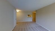 This single-floor space features an open living area finished with wall-to-wall carpeting in a neutral beige tone, complementing the light-colored walls and white baseboards throughout. The room offers a generous sense of space, providing ample room for a variety of furniture arrangements. A flush-mount ceiling light fixture illuminates the area with a warm glow, contributing to the overall inviting ambiance. A notable architectural detail is the pass-through window cut into the wall, which connects the living area to the adjacent kitchen, allowing light and visibility to flow between the two spaces. The kitchen area, visible through the pass-through, features what appears to be countertop surfaces and cabinetry. A white-paneled door on the left side of the living area provides access to an additional room or utility space. The open entryway on the right side of the space leads further into the unit, creating a sense of connectivity between rooms. The overall design is clean and straightforward, with neutral finishes throughout that provide a versatile backdrop suitable for a range of interior styles.