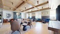 Located at 1002 N Fourth St in Flagstaff, Arizona, this single-floor living lounge is a thoughtfully designed communal space that blends contemporary style with warm, lodge-inspired elements. The room is anchored by a striking stacked-stone fireplace with a built-in linear firebox, a wall-mounted television, and dark open shelving accented with curated décor. Coffered wood-beam ceilings add architectural depth and a sense of warmth overhead, while light hardwood flooring runs throughout, grounding the space in a clean, natural aesthetic. Large windows dressed in rich navy blue curtains allow abundant natural light to fill the room, framing views of the surrounding building exterior. Seating is arranged in thoughtful groupings, including a generous neutral sectional sofa, a pair of sculptural brown leather wingback chairs, and a cozy banquette dining nook with a stone accent wall. A blue cabinetry unit with a built-in wine cooler serves as both a functional and decorative focal point. Textured area rugs define each seating zone, while track lighting and flush-mount ceiling fixtures provide layered illumination. The overall ambiance is sophisticated yet comfortable, making this lounge an ideal gathering and relaxation space.