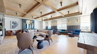 Located at 1002 N Fourth St in Flagstaff, Arizona, this single-floor living lounge is a thoughtfully designed communal space that blends contemporary style with warm, lodge-inspired elements. The room is anchored by a striking stacked-stone fireplace with a built-in linear firebox, a wall-mounted television, and dark open shelving accented with curated décor. Coffered wood-beam ceilings add architectural depth and a sense of warmth overhead, while light hardwood flooring runs throughout, grounding the space in a clean, natural aesthetic. Large windows dressed in rich navy blue curtains allow abundant natural light to fill the room, framing views of the surrounding building exterior. Seating is arranged in thoughtful groupings, including a generous neutral sectional sofa, a pair of sculptural brown leather wingback chairs, and a cozy banquette dining nook with a stone accent wall. A blue cabinetry unit with a built-in wine cooler serves as both a functional and decorative focal point. Textured area rugs define each seating zone, while track lighting and flush-mount ceiling fixtures provide layered illumination. The overall ambiance is sophisticated yet comfortable, making this lounge an ideal gathering and relaxation space.