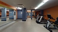 This fitness center in Westlake, Ohio is a well-equipped, single-floor facility designed to support a comprehensive workout experience. The space is defined by a bold color palette of deep navy blue and energizing orange, applied across structural columns and accent walls, creating a dynamic and motivating atmosphere throughout. Durable speckled flooring covers the entire area, providing both safety and practicality underfoot.

The layout is thoughtfully organized into distinct workout zones. The cardio area features multiple treadmills, elliptical trainers, and stair-climber machines, including Spirit and Precor branded equipment, arranged to allow ample space between users. A dedicated free weights section houses a full rack of dumbbells alongside a weight bench, accompanied by motivational wall decals that reinforce a goal-oriented mindset. A mirrored wall in the strength training alcove enhances the sense of space while allowing users to monitor their form during barbell and bench press exercises.

Additional strength training options include a cable machine station, supporting a variety of resistance-based exercises. The space is lit by fluorescent overhead fixtures mounted to a textured ceiling, maintaining consistent visibility throughout. Wall-mounted mirrors and a flat-screen television add functional and recreational elements to the environment. The facility is accessible via a standard entry door and is designed to serve users of varying fitness levels.