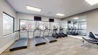 This fitness center in Memphis, TN, is a well-appointed, single-level exercise facility designed to support a well-rounded workout routine. The space features neutral gray walls, textured carpet tile flooring, and a clean, modern aesthetic throughout. Recessed panel lighting in the ceiling ensures consistent, bright illumination across the entire floor, complementing the natural light that streams in through multiple large windows along the exterior walls. These windows provide views of the surrounding tree-lined outdoor environment, creating an open, airy atmosphere despite the room's compact footprint.

The cardio zone is a focal point of the space, lined with treadmills and elliptical trainers positioned to face the windows. Two wall-mounted flat-screen televisions are installed above the machines, offering entertainment during workouts. A full-length mirror spans one wall, visually expanding the room and allowing users to monitor their form.

On the strength training side, a multi-station cable machine and weight bench are available, complemented by stability balls stored in the corner. The space also includes a stationary bike, rounding out the variety of available equipment. A dedicated entry door provides direct exterior access, adding convenience and functionality to this efficient, purpose-built fitness facility.