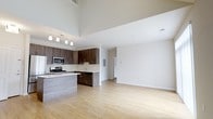 This contemporary two-floor space is defined by its striking open-concept design and impressive soaring ceilings that lend an expansive, airy quality to the main living area. Light-toned hardwood flooring flows seamlessly throughout, enhancing the sense of openness and cohesion.

The kitchen is a standout feature, outfitted with rich dark-wood cabinetry, light-toned countertops, and a full suite of stainless steel appliances, including a gas range, microwave, and dishwasher. A peninsula with a clean-lined finish provides additional counter space and serves as a natural divider between the kitchen and the generous living area. Pendant track lighting above the kitchen adds both function and a modern aesthetic accent.

The open living area benefits from the dramatic ceiling height, creating a grand atmosphere ideal for both everyday living and entertaining. Crisp white walls and clean architectural lines contribute to the bright, minimalist character of the space. A dedicated utility panel is thoughtfully integrated along one wall, maintaining the streamlined aesthetic. With two floors, the space offers versatile living arrangements and ample room for a variety of lifestyles. Overall, the design balances modern sophistication with practical functionality.
