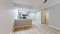 This single-floor unit showcases a clean, modern aesthetic highlighted by light wood-look flooring that runs continuously throughout the space, creating a cohesive and airy feel. The open-concept kitchen is a standout feature, anchored by a peninsula with a white countertop and integrated sink, offering both workspace and a natural divider between the kitchen and living areas. Gray shaker-style cabinetry lines the kitchen walls, complemented by stainless steel appliances including a refrigerator, electric range, and over-the-range microwave. A tiled backsplash and recessed lighting add refined detail to the cooking space. The living area is generously sized with a ceiling fan overhead and access to additional rooms through a paneled interior door. The bedrooms continue the light flooring theme and feature textured stucco-style accent walls that add visual depth and character. One bedroom includes a ceiling fan and a window fitted with horizontal blinds, allowing for natural light control. Throughout the unit, smooth gray walls, white baseboards, and textured ceilings contribute to a neutral, move-in-ready palette. Ceiling-mounted HVAC vents are present in multiple rooms, ensuring consistent climate control. Overall, the space presents a fresh, updated interior with functional design elements suited for comfortable everyday living.