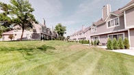 This single-floor residence is part of a well-maintained multi-unit community characterized by classic architectural design. The exteriors feature neutral-toned vinyl siding, pitched rooflines with prominent chimneys, and neatly landscaped surroundings with manicured lawns, ornamental shrubs, and mature trees — creating a peaceful, park-like setting shared across the community.

Inside, the open-concept layout connects the kitchen and living areas seamlessly, enhanced by soaring vaulted ceilings that lend a sense of spaciousness and airiness. A clerestory window positioned high on the wall draws in natural light, brightening the main living space. Durable wood-look flooring runs throughout the common areas, complementing the warm interior palette.

The kitchen is a functional highlight, outfitted with rich honey-toned wood cabinetry, light granite-style countertops, and a double stainless steel sink. Black appliances — including an electric range, over-the-range microwave, dishwasher, and refrigerator — provide a cohesive, modern look. Ample upper and lower cabinet storage ensures excellent utility. The kitchen opens directly toward the entryway and living area, making the space ideal for everyday living and entertaining alike.
