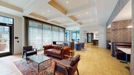 Located at 50 S Grant Ave in downtown Columbus, Ohio, Library Park's amenity floor is a thoughtfully designed communal space that blends classic sophistication with modern comfort. The main lounge area is anchored by a tufted leather Chesterfield sofa, mid-century modern armchairs, and Persian-style area rugs atop herringbone-patterned hardwood flooring. Coffered ceilings with recessed lighting add architectural depth, while floor-to-ceiling windows invite abundant natural light and views of the surrounding streetscape. Sheer drapery panels soften the generous glazing throughout. A centrally positioned bar features an expansive white quartz countertop, dark cabinetry, built-in stainless steel refrigeration units, and gold pendant lighting over glass-front upper cabinets — ideal for entertaining. Booth-style banquette seating with richly textured wall coverings provides an intimate dining alcove. A private conference or dining room offers a moody, refined atmosphere with deep navy walls, a statement globe chandelier, a substantial dark wood table, and plush red velvet seating. The entry area features hexagonal tile flooring and a seating nook with blue barrel chairs, welcoming residents and guests into this well-appointed gathering space.