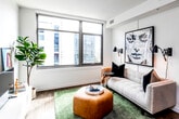 This single-floor residence offers a thoughtfully designed layout with a contemporary aesthetic that flows seamlessly from room to room. The living area serves as the heart of the space, featuring large picture windows that frame urban cityscape views and fill the room with natural light. Warm wood-tone flooring runs throughout, complementing the clean white walls and modern decor. A textured accent wall provides visual interest behind the media console, which anchors one side of the living space.

The kitchen is a standout feature, outfitted with sleek dark cabinetry, stainless steel appliances including a full-size refrigerator, electric range, microwave, and dishwasher. A white subway tile backsplash adds a classic touch, while open shelving provides practical storage and display space. Under-cabinet LED strip lighting enhances the modern feel, and a kitchen island offers additional prep and dining space.

The bedroom is tranquil and well-proportioned, featuring a closet with sliding doors and built-in shelving. The attached bathroom includes a full soaking tub and shower combination with white tile surround, dark hexagonal floor tile, and a single vanity. Throughout, the space maintains a cohesive, modern residential character with clean lines and quality finishes.