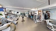 Located in the heart of downtown Montréal on Rue de Bleury, this fitness center offers a comprehensive and well-organized workout environment within a bright, single-floor layout. Large floor-to-ceiling windows line one side of the space, flooding the gym with abundant natural light while providing views of the surrounding urban cityscape — an energizing backdrop for any workout session.

The gym is thoughtfully divided into dedicated zones to accommodate a wide variety of training styles. The cardio area features multiple treadmills, elliptical trainers, a stair climber, and rowing machines, all positioned to take advantage of the natural light and city views. Full-length mirrors along the walls visually expand the space and assist with form during exercise.

The strength training section is equally well-appointed, with a multi-station cable machine, weight benches, a dedicated free weights rack offering a full range of dumbbells, and a hanging heavy punching bag for boxing-style training. Stability balls and additional accessories round out the functional fitness offerings.

Durable rubber flooring provides safety and comfort throughout the space, while recessed and linear overhead lighting ensures consistent illumination. A wall-mounted flat-screen television adds convenience for entertainment during workouts. The clean, modern aesthetic — characterized by neutral tones and an uncluttered layout — creates an efficient and motivating environment for users of all fitness levels.