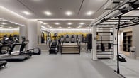 This single-floor fitness center is designed with a clean, contemporary aesthetic and thoughtfully organized into distinct training zones. The space is finished throughout with smooth gray flooring and neutral gray walls, creating a cohesive, professional environment. Recessed ceiling lighting, complemented by linear LED cove lighting, ensures the entire facility is evenly and brightly illuminated.

The cardio zone is well-appointed with a row of treadmills, ellipticals, spinning bikes, rowing machines, and stair climbers. A large wall-spanning mirror in this section visually expands the space and is topped by a striking bold yellow and white graphic mural featuring motivational text, serving as a defining design feature of the facility.

A dedicated functional training area houses a ceiling-mounted rig system supporting a heavy punching bag, suspension trainers, and various attachments. A Precor cable machine and additional strength equipment are positioned nearby for targeted resistance training.

The strength and multi-use zone provides ample open floor space alongside weight benches, cable systems, and a stairmill. Framed glass entry doors with an adjacent planter provide an elegant transition into the space. Throughout, the layout prioritizes clear circulation paths and a streamlined, upscale fitness environment.