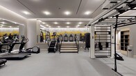 This single-floor fitness center is designed with a clean, contemporary aesthetic and thoughtfully organized into distinct training zones. The space is finished throughout with smooth gray flooring and neutral gray walls, creating a cohesive, professional environment. Recessed ceiling lighting, complemented by linear LED cove lighting, ensures the entire facility is evenly and brightly illuminated.

The cardio zone is well-appointed with a row of treadmills, ellipticals, spinning bikes, rowing machines, and stair climbers. A large wall-spanning mirror in this section visually expands the space and is topped by a striking bold yellow and white graphic mural featuring motivational text, serving as a defining design feature of the facility.

A dedicated functional training area houses a ceiling-mounted rig system supporting a heavy punching bag, suspension trainers, and various attachments. A Precor cable machine and additional strength equipment are positioned nearby for targeted resistance training.

The strength and multi-use zone provides ample open floor space alongside weight benches, cable systems, and a stairmill. Framed glass entry doors with an adjacent planter provide an elegant transition into the space. Throughout, the layout prioritizes clear circulation paths and a streamlined, upscale fitness environment.