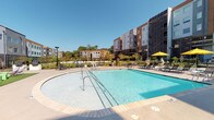 Located at 125 Anderson Hwy in Clemson, South Carolina, this contemporary residential community showcases a bold, modern architectural design characterized by multi-story buildings with eye-catching facades that blend gray, orange, and wood-tone panel accents. The exterior design reflects a fresh, urban aesthetic well-suited to the Clemson area.

The outdoor amenity spaces are a standout feature of this property. A generously sized resort-style swimming pool serves as the centerpiece of the community, offering both a rounded shallow entry area and a larger rectangular section, surrounded by a spacious concrete pool deck furnished with lounge chairs and bright yellow umbrellas. The pool area is framed by lush landscaping, iron fencing, and views of the surrounding buildings and tree line.

Adjacent to the pool, a lively courtyard area provides multiple outdoor gathering spaces, including vibrant orange canopy cabanas with lounge seating, turf areas with colorful modular seating, and patio dining tables perfect for outdoor meals. A separate intimate lounge nook features white outdoor sofas, armchairs, and a green accent table shaded by a yellow umbrella, creating a relaxed social atmosphere.

The community's outdoor spaces are thoughtfully designed to encourage interaction and leisure, offering a variety of settings suited for relaxation and socializing under the open South Carolina sky.