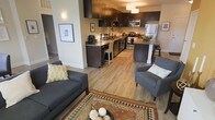 This single-floor residence showcases a thoughtfully designed open-concept layout that seamlessly connects the living and kitchen areas into one cohesive, functional space. The living area is anchored by a comfortable grey sofa and matching armchair, accented with decorative throw pillows and a vibrant patterned area rug that adds warmth and character to the light wood flooring beneath.

The kitchen is a standout feature, outfitted with rich dark cabinetry that contrasts beautifully against light countertops and stainless steel appliances, including a refrigerator, range, and built-in microwave. A kitchen island with bar seating provides a casual dining option and doubles as additional prep space. Pendant lighting overhead adds both task illumination and a modern design touch.

The light hardwood-style flooring extends throughout, creating a sense of continuity and openness across the entire floor plan. Neutral wall tones complement the dark cabinetry and contemporary furnishings, while wall-mounted lighting fixtures contribute to a warm, inviting ambiance. The open flow between spaces makes the layout feel spacious and airy, ideal for both everyday living and entertaining.