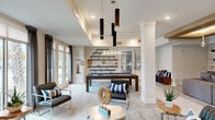 This thoughtfully designed clubhouse in Cayce, South Carolina offers a seamless blend of social and leisure spaces across a single floor. The main lounge area makes an immediate impression with soaring ceilings, dramatic pendant lighting, and an open layout anchored by a full-sized pool table. Stylish seating arrangements, including sleek chrome-frame chairs and plush sofas, are accented with blue and white throw pillows, creating a contemporary and inviting atmosphere. Large floor-to-ceiling windows flood the space with natural light and offer views of the outdoor pool area, complete with lounge chairs and lush landscaping. The kitchen is a standout feature, boasting warm wood cabinetry, a striking herringbone wood-paneled accent wall with mirrored inlays, marble-veined countertops, and stainless steel appliances. A dedicated dining area with a rustic wood farmhouse table, tufted upholstered chairs, and an elegant glass chandelier sits beneath a geometric coffered ceiling detail. The media lounge, slightly set apart from the main area, features a tray ceiling with wood accents, built-in shelving, and a wall-mounted flat-screen television, offering a quieter retreat within the clubhouse. Tile flooring and plush carpeting distinguish the various zones, while curated décor and art pieces throughout add a polished, cohesive finishing touch.