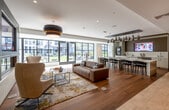This thoughtfully designed single-floor amenity space offers a seamless blend of comfort and contemporary style across several distinct yet open-plan zones. The entry is framed by full-height glass doors, immediately revealing the expansive, light-filled interior beyond.

Rich hardwood flooring flows throughout, anchoring a central lounge area furnished with plush sofas, accent chairs, and an abstract area rug. A sleek, linear gas fireplace is integrated into a bold dark feature wall, accompanied by a large wall-mounted television — creating an inviting focal point for relaxation. Adjacent private booth seating provides a more intimate setting for smaller gatherings.

A freestanding double-sided fireplace with a stone surround divides the space, separating the lounge from a casual dining nook centered beneath an elegant tiered chandelier. Black swivel chairs and a round table complete this area.

The kitchenette runs along one wall, outfitted with white cabinetry, quartz countertops, a commercial coffee machine, under-counter refrigeration, and a wall-mounted display. A long communal dining table with pendant lighting overhead serves as an additional gathering hub.

Floor-to-ceiling steel-framed windows and glass doors along the perimeter flood the space with natural light while offering direct views of and access to an outdoor courtyard, seamlessly connecting interior and exterior living.