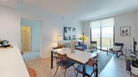 This contemporary single-floor residence is designed with a clean, modern aesthetic and an open-concept layout that seamlessly connects the living, dining, and kitchen areas. Light wood-tone flooring flows throughout the main living spaces, complementing the bright white walls and recessed lighting overhead.

The living area is anchored by floor-to-ceiling windows that flood the space with natural light and offer expansive views of the surrounding cityscape and sky. A glass-top coffee table and stylish seating arrangements make this an inviting area for relaxation and entertaining.

The dining area features a marble-topped table with mixed seating, positioned to take advantage of the natural light and views. The kitchen is outfitted with crisp white cabinetry, a large quartz waterfall island with bar seating for multiple guests, and a full suite of stainless steel appliances including a refrigerator, microwave, and range. A stacked washer and dryer unit is conveniently tucked adjacent to the kitchen.

The primary bedroom offers plush carpeting, framed wall art, and direct access to an en-suite bathroom, along with a dedicated closet space. The second bedroom is equally well-appointed, featuring soft carpeting and access to a full bathroom complete with a soaking tub and shower combination. Throughout, the space maintains a cohesive, contemporary feel with thoughtful finishes and functional design.