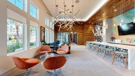 Located on International Drive in Orlando, Florida, this contemporary community clubhouse delivers a sophisticated and vibrant atmosphere across a single, generously proportioned floor. The main lounge area immediately captivates with soaring double-height ceilings adorned by a striking sculptural chandelier composed of intersecting linear light fixtures. Warm, richly textured wood-paneled accent walls contrast elegantly with crisp white surfaces, while floor-to-ceiling windows flood the space with abundant natural light and offer views of the surrounding outdoor greenery. A long marble-topped bar counter with teal upholstered barstools anchors one side of the main space, complemented by a mounted flat-screen TV and a vibrant mosaic art installation as a backdrop. The lounge features eclectic, colorful seating arrangements, including retro-style swivel chairs and modular sectional pieces, alongside a game area outfitted with foosball and shuffleboard tables and bold large-scale wall art. A quieter reading nook with built-in bookshelves and a glass-partitioned lounge area provide more intimate settings. The dedicated office features a built-in white desk set against a distinctive teal textured accent wall, offering a focused workspace. A connecting hallway leads to four well-appointed bathrooms, completing this thoughtfully designed facility.