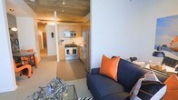 This single-floor residence showcases a thoughtfully designed open-concept layout that seamlessly blends industrial and contemporary aesthetics. Exposed concrete ceilings with track lighting lend a loft-like character throughout the space, creating a distinctive urban atmosphere.

The living area is anchored by a sleek, low-profile sofa in deep navy, accented with vibrant orange cushions that introduce a bold pop of color. A geometric glass coffee table adds a modern, artistic touch, while the light hardwood flooring ties the open space together with warmth and continuity.

The kitchen flows directly from the living area and is outfitted with stainless steel appliances, including a gas range, built-in microwave, and a full-size refrigerator. Light wood cabinetry provides ample storage while keeping the space feeling open and airy. Clean countertops and an efficient layout make it both functional and visually appealing.

A hallway visible from the main living space suggests additional rooms beyond, maintaining a sense of depth and separation within the residence. Neutral wall tones throughout allow the architectural details and curated furnishings to take center stage. Overall, this space combines industrial character with modern comfort, creating a dynamic and inviting environment.