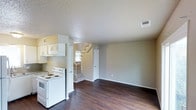 This two-story space offers a practical and straightforward layout across its main and upper levels. The main floor presents an open concept design where the kitchen and living area flow seamlessly together. The kitchen is equipped with white cabinetry mounted above the cooking area, providing functional storage, alongside a white electric range and an overhead ventilation unit. The flooring throughout the main level features dark hardwood-style planks that add warmth and contrast against the neutral, light-toned walls. A textured ceiling adds subtle visual detail overhead, while a ceiling-mounted HVAC vent ensures climate control throughout the space. Natural light filters in through a window near the staircase, brightening the transition area between floors. The staircase, carpeted in a neutral tone, connects the main level to the upper floor, accompanied by a simple railing. A closed door near the base of the stairs likely leads to additional storage or a utility space. The living area is open and unobstructed, offering flexibility for various furniture arrangements. Overall, the space maintains a clean, functional aesthetic with a neutral palette throughout.