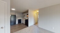 This single-floor unit is designed with a practical, straightforward layout that maximizes the use of available space. The kitchen serves as a functional hub, outfitted with white cabinetry extending from countertop to ceiling, light granite-style countertops, and a full suite of black appliances including a gas range, dishwasher, refrigerator, and an under-cabinet range hood. A stainless steel sink is centrally positioned, and wood-look flooring runs throughout the kitchen and entry areas, providing durability and easy maintenance.

The living and bedroom areas are finished with wall-to-wall carpet in a neutral tone, contributing to a quieter, comfortable atmosphere. The living space is generously proportioned with baseboard heating units along the walls and multiple electrical outlets. A bedroom features a large window that allows natural light to fill the room, offering views of surrounding greenery. Closet doors provide convenient storage directly off the main living area.

The bathroom is clean and well-appointed, featuring a tub-shower combination with a decorative tile accent border, a vanity with white cabinetry, a toilet, and wood-look flooring. Throughout the unit, neutral gray walls, textured ceilings, and consistent trim work create a cohesive, understated aesthetic.