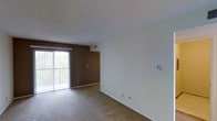 This single-floor residential space offers a clean and functional layout designed for comfortable everyday living. The main living area is finished with wall-to-wall carpeting in a warm neutral tone, creating a cozy and inviting atmosphere. A large sliding glass door floods the room with natural light and provides direct access to an outdoor balcony, offering views of surrounding greenery. A contrasting dark brown accent wall frames the sliding door, adding a touch of visual interest to the otherwise bright and neutral interior. The textured ceiling and crisp white walls contribute to the space's understated, move-in-ready aesthetic. A wall-mounted thermostat is conveniently located for easy climate control. The open, unobstructed floor plan allows for versatile furniture arrangement to suit a variety of lifestyles. Ample wall space provides flexibility for customization and personalization. Overall, the space is characterized by its simplicity, practicality, and the natural light that defines its welcoming ambiance.