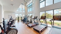 Located at Patriots Point in Mount Pleasant, South Carolina, this two-floor fitness facility offers a well-appointed and light-filled environment designed for a comprehensive workout experience. The ground floor serves as the primary cardio zone, showcasing soaring double-height ceilings framed by floor-to-ceiling glass walls that flood the space with natural light and provide views of the surrounding landscape. Light hardwood flooring runs throughout, complementing the bright white walls and creating a clean, modern aesthetic. A row of commercial-grade treadmills, ellipticals, stair climbers, and stationary bikes — including Peloton cycles — are thoughtfully arranged to take full advantage of the outdoor views. A dedicated glass-enclosed studio space on this level features mirrored walls and hardwood floors, ideal for group fitness, yoga, or personal training sessions. Exposed light brick accent walls add a warm, textural contrast to the otherwise contemporary design. The upper floor transitions to a strength training zone, carpeted for comfort and equipped with a full array of cable machines, weight stations, and benches. A glass-railed staircase connects the two levels, maintaining visual openness throughout. The facility also includes restroom access, built-in lockers, and a double-door entrance with a dramatic two-story glazed entryway.