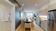 This single-floor residence showcases a thoughtfully designed open-plan layout where the kitchen, dining, and living areas flow seamlessly together, unified by light hardwood flooring and recessed lighting throughout. The galley-style kitchen is outfitted with stainless steel appliances—including a range, microwave, dishwasher, and refrigerator—complemented by flat-front cabinetry in warm wood tones, under-cabinet lighting, and a sleek marble-look backsplash with white quartz countertops. A generously sized dining table anchors the central space, flanked by upholstered chairs, while a deep teal accent wall adds bold contrast to the otherwise neutral palette.

The living area is positioned toward the front of the space, anchored by floor-to-ceiling windows that frame urban cityscape views and flood the room with natural light. A ceiling fan provides additional comfort in both the living and bedroom areas.

The bedroom features a striking deep blue accent wall adorned with geometric wall art, a low-profile platform bed, and a tall vertical window that continues the city view theme. The bathroom is finished in full marble-look tile—covering both floors and walls—with a frameless glass-enclosed walk-in shower, a floating wood vanity, and an illuminated mirror, creating a spa-like atmosphere.