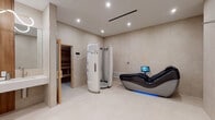 This single-level wellness space is thoughtfully designed as a comprehensive personal spa and recovery suite. The interior is finished throughout in warm beige large-format floor tiles and smooth plaster-toned walls, creating a clean, spa-like aesthetic that feels both calming and clinical.

The main treatment area serves as the focal point of the suite, housing a full-length vertical cryotherapy or tanning pod alongside a high-tech zero-gravity massage chair equipped with its own integrated touchscreen display. Recessed ceiling lighting and a linear air diffuser overhead lend the space a polished, modern finish.

Adjacent to the main area, a traditional cedar-lined infrared sauna is accessible through a wood-framed glass door, offering a warm contrast to the room's neutral palette. A dedicated vanity station with dual sinks, warm wood cabinetry surrounds, and backlit mirrors with decorative lighting provides a refined grooming area nearby.

The suite also includes a walk-in shower finished entirely in matching large-format beige tiles, outfitted with a sleek stainless steel shower panel featuring multiple controls and a handheld fixture. A separate doorway leads to an additional relaxation area, complete with what appears to be a thermal or infrared body wrap mat and a small wooden bench. The overall ambiance is serene, cohesive, and purpose-built for wellness.