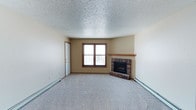 This single-floor residence offers a straightforward and functional layout designed for comfortable everyday living. The living area serves as the centerpiece of the space, characterized by soft wall-to-wall carpeting underfoot and a classic textured popcorn ceiling above. A brick corner fireplace with a wooden mantel provides a warm focal point, adding both visual interest and a cozy ambiance to the room. Natural light filters in through a double-pane window framed in warm wood tones, offering a view to the outdoors. Baseboard heaters line the perimeter of the living area, ensuring efficient climate control throughout the seasons. The neutral beige walls create a versatile backdrop, easily adaptable to a variety of interior styles. A doorway connects the living area to additional spaces within the home, maintaining a smooth flow throughout the floor plan. Overall, the space presents a clean, move-in ready environment with practical features that prioritize comfort and functionality.