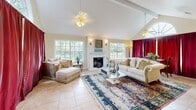 Located at 1331 S Harvey Mitchell Parkway in College Station, TX, this single-room living space makes a strong architectural impression with its soaring vaulted ceiling accented by exposed beams and dual ceiling fans. The room is generously filled with natural light, courtesy of multiple large windows along the walls and a striking arched transom window that serves as a focal point, flooding the space with warm, even illumination throughout the day.

At the heart of the room stands a white marble fireplace with an ornate surround and mantle, flanked by decorative wall sconces that add a warm, classic ambiance. Light-toned ceramic tile flooring runs throughout, complemented by a richly patterned area rug that anchors the seating arrangement. The living area comfortably accommodates a classic sofa, chaise lounge, and a glass-top coffee table, creating an inviting and functional layout.

A set of white French doors leads to an adjoining area, while dramatic floor-length crimson curtains add a bold contrast to the room's otherwise soft, neutral palette. A wall-mounted television and console area provide a modern, practical touch. The combination of architectural detail, ample natural light, and elegant finishes gives this living space a timeless, refined character.