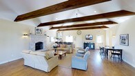 This generously sized living space is defined by its soaring vaulted ceilings adorned with rich, dark-stained exposed wooden beams that draw the eye upward and lend a warm, rustic character to the room. The hardwood floors throughout add natural warmth and continuity, complementing the overall coastal-inspired aesthetic.

A striking stone fireplace serves as a prominent focal point along one wall, framed by a classic white mantel and providing a welcoming anchor to the main seating area. The living space is furnished with a collection of comfortable sofas and armchairs in soft, neutral tones, arranged to encourage conversation and relaxation. A wooden coffee table grounds the central seating arrangement atop a large area rug.

Natural light filters in through multiple windows and a glass-paneled door, brightening the cream-colored walls. Pendant lighting and recessed fixtures supplement the natural illumination. A wall-mounted flat-screen television rests on a dark media console, while a decorative wall clock and framed nautical artwork reinforce the space's coastal charm. A dining area with bar-height seating is visible toward the back of the room.
