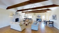 This generously sized living space is defined by its soaring vaulted ceilings adorned with rich, dark-stained exposed wooden beams that draw the eye upward and lend a warm, rustic character to the room. The hardwood floors throughout add natural warmth and continuity, complementing the overall coastal-inspired aesthetic.

A striking stone fireplace serves as a prominent focal point along one wall, framed by a classic white mantel and providing a welcoming anchor to the main seating area. The living space is furnished with a collection of comfortable sofas and armchairs in soft, neutral tones, arranged to encourage conversation and relaxation. A wooden coffee table grounds the central seating arrangement atop a large area rug.

Natural light filters in through multiple windows and a glass-paneled door, brightening the cream-colored walls. Pendant lighting and recessed fixtures supplement the natural illumination. A wall-mounted flat-screen television rests on a dark media console, while a decorative wall clock and framed nautical artwork reinforce the space's coastal charm. A dining area with bar-height seating is visible toward the back of the room.