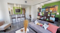 Located in the heart of downtown Jacksonville, Florida, this single-floor unit offers a thoughtfully designed open-concept layout with a contemporary urban aesthetic. The living area is anchored by polished concrete floors, large windows framing sweeping city skyline views, and neutral walls accented by bold green feature walls that add a vibrant pop of color. A stylish open shelving unit serves as a functional room divider, adding character and display space to the living area.

The kitchen features light maple cabinetry, laminate countertops, and black appliances including an electric range with overhead microwave. A breakfast bar with bar-height seating creates a seamless transition between the kitchen and living space, making it ideal for casual dining and entertaining.

The bathroom is clean and well-appointed, with a single-sink vanity topped with a white countertop, warm wood cabinetry, a large mirror with multi-bulb vanity lighting, and a shower/tub combination. The floor plan, visible in the dollhouse view, reveals an efficient use of space with clearly defined areas for living, dining, cooking, and sleeping — all connected in a cohesive, modern design that makes the most of its downtown location.