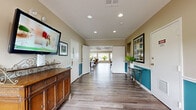 Located at 5001 SW 20th St in Ocala, Florida, this single-floor community clubhouse is designed to foster comfort, socializing, and relaxation. The entrance corridor sets a warm tone with rich wood-look flooring, crown molding, teal accent wainscoting, and framed artwork lining the walls, leading visitors naturally into the heart of the space.

The main gathering area is expansive and open, combining a lounge and dining zone seamlessly. Multiple round dining tables with wooden chairs are arranged throughout, encouraging communal interaction. A standout brick fireplace with a classic white mantel anchors one end of the room, adding warmth and character. Adjacent seating areas with comfortable upholstered furniture provide relaxed conversation spaces.

A wall of large picture windows floods the interior with abundant natural light and offers sweeping views of the outdoor patio, pool area, and lush Florida palm trees beyond. The bright, airy ambiance is further enhanced by recessed lighting and a coffered ceiling detail overhead.

The kitchenette area is sleek and functional, featuring modern gray cabinetry, a white island with bar seating, stainless steel appliances, and floor-to-ceiling mirrored panels that amplify the sense of space. Throughout the facility, consistent wood-look flooring and a neutral warm color palette create a cohesive, inviting atmosphere ideal for community gatherings.
