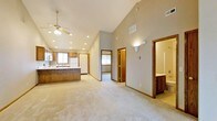 This single-story home showcases a thoughtfully designed open-concept floor plan that seamlessly connects the main living area with the kitchen. The space is defined by striking vaulted ceilings that rise dramatically overhead, enhancing the sense of openness and volume throughout. Recessed lighting and a ceiling fan with a light fixture provide practical illumination while complementing the airy feel of the main area.

The kitchen features warm wood cabinetry, a peninsula with an integrated sink, and white appliances, including a refrigerator. A window above the sink allows natural light to filter in. The flooring transitions between the kitchen and the carpeted living areas, which are finished in neutral tones that contribute to the home's understated, welcoming ambiance.

Neutral gray-toned walls are trimmed with warm wood door frames, adding a cohesive design detail throughout. Multiple doorways lead to additional rooms, including what appears to be a bedroom with its own window and a bathroom featuring a vanity with wood cabinetry. The layout is practical and well-organized, making efficient use of the available space on a single level.