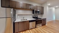 This thoughtfully designed single-floor unit blends contemporary finishes with functional living spaces throughout. The kitchen is a standout feature, outfitted with dark espresso cabinetry extending to the ceiling, granite-look countertops, a classic white subway tile backsplash, and a full suite of stainless steel appliances including a range, microwave, dishwasher, and refrigerator. Warm, light-toned hardwood-style flooring runs throughout the main living and kitchen areas, lending a cohesive and inviting feel. The bathroom continues this aesthetic with the same warm flooring, a dark wood vanity with ample drawer storage, a single-basin sink with a large mirror above, a wall-mounted light bar, and a full soaking tub with tiled surround. A tall storage cabinet provides additional organization in the bathroom. The bedroom features plush carpet underfoot and is bathed in natural light from a large, floor-to-ceiling window that frames views of outdoor greenery. The open layout, visible in the overhead dollhouse view, reveals generous built-in shelving and storage throughout, making efficient use of the available space. Clean white walls and neutral tones throughout create a bright, modern ambiance.