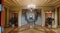 This distinguished foyer makes an immediate impression with its grand architectural design and refined classical aesthetic. At its centerpiece, a pair of symmetrical curved staircases sweep upward on either side, adorned with gilded balustrades and detailed newel posts that reflect exceptional craftsmanship. Flanking the staircases, tall fluted pilasters with decorative capitals frame the space, reinforcing the formal, stately character of the interior.

Above, a multi-arm crystal and brass chandelier casts warm, ambient light across the room, complementing the ornate coffered ceiling with its subtle molding details. The flooring is a polished inlaid design featuring geometric patterns in contrasting tones, adding visual depth and grandeur underfoot.

A long corridor extends beyond the staircase arch, drawing the eye deeper into the space and hinting at the scale of the building. On either side of the foyer, glazed wooden doors with divided-light panes provide access to adjacent rooms, maintaining an open yet structured flow. The overall ambiance is one of timeless elegance, combining classical architectural elements with luxurious finishes to create a commanding and welcoming entrance.