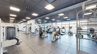 Located at 221 Harborside Dr in Schenectady, NY, this modern fitness facility spans a generous single floor and is divided into two distinct gym areas, each thoughtfully designed to support a wide range of workout needs. The main gym area is bright and open, benefiting from expansive floor-to-ceiling windows that flood the space with natural light and offer views of the surrounding neighborhood. It features a comprehensive selection of True-brand cardio equipment—including treadmills, ellipticals, and stationary bikes—alongside a robust selection of strength machines, free weights, a power rack, and benches arranged across durable large-format tile flooring. Ceiling fans and recessed panel lighting maintain a comfortable and well-lit environment throughout. The second gym area functions as a dedicated studio and functional training space, lined with full-length mirrors along the walls to enhance spatial awareness during workouts. This room is equipped with exercise mats, stability balls, medicine balls, resistance bands, a Pilates reformer, plyo boxes, and additional cardio equipment, making it ideal for group classes, personal training, or independent functional fitness sessions. A connecting corridor with additional specialty equipment, including a stair-climbing machine, links the two areas seamlessly. Together, the two gym spaces create a comprehensive, professional-grade fitness environment suited for diverse training modalities.