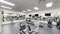 This fitness center offers a comprehensive workout environment designed to accommodate a wide range of exercise needs. The space is finished with a durable rubber flooring throughout, providing both safety and comfort during training sessions. A dropped ceiling with recessed panel lighting ensures the room is evenly and brightly illuminated, contributing to an open, energetic atmosphere.

Large wall-to-wall mirrors line multiple walls, visually expanding the space while allowing users to monitor their form during exercise. Multiple flat-screen televisions are mounted above the mirrors, providing entertainment options during cardio sessions.

The cardio zone features a row of treadmills and elliptical trainers from reputable brands including True and Hoist. The strength training area is equally well-appointed, housing a Hoist functional trainer cable machine, a Smith machine, and a dedicated free weights section. The free weight area includes a full rack of kettlebells, dumbbells, and medicine balls in various sizes, along with resistance bands and exercise mats. Workout reference charts are posted on the walls for user guidance.

Overall, the fitness center presents a clean, organized, and professional environment well-suited for both cardio and strength training routines.