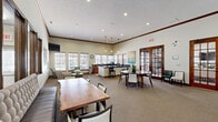 Located at 1021 Stoneridge Dr in Seymour, Indiana, this single-floor community space combines a fully equipped kitchen and an open living area into one cohesive, welcoming environment. The layout makes efficient use of its footprint, offering multiple distinct zones for gathering, dining, and relaxing.

The kitchen is appointed with warm honey-toned wood cabinetry, dark granite countertops, and tile flooring. A wraparound counter configuration provides generous workspace, while a central island with bar seating serves as a casual gathering point. Pendant lights hang above the island, and large windows flood the area with natural light through slatted wood blinds.

The adjoining living and dining area features soaring ceilings with elegant crown molding and recessed lighting, creating an airy, open atmosphere. Multiple seating arrangements accommodate various group sizes — from intimate lounge clusters with upholstered armchairs and a round coffee table, to longer dining tables with mixed wood and upholstered chairs. A wall-mounted flat-screen television anchors one corner of the lounge area. French doors on one wall open to an outdoor view, enhancing the sense of connectivity with the surrounding environment. Patterned carpet throughout ties the space together with a clean, cohesive aesthetic.