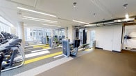 This two-floor fitness facility offers a thoughtfully designed environment for health and wellness. The gym floor is immediately striking, featuring a bold patterned carpet with yellow, white, and grey geometric striping that adds energy and visual direction to the space. A comprehensive array of strength training and cable machines are arranged throughout the floor, providing users with a wide variety of workout options. Large windows line the perimeter walls, flooding the interior with abundant natural light and offering views of the surrounding outdoor environment, creating an open and motivating atmosphere. The ceiling is finished with clean white paneling and fitted with long fluorescent strip lighting, ensuring consistent, bright illumination throughout. Exposed industrial-style ductwork adds a modern, urban aesthetic to the otherwise clean design. Structural columns are distributed across the floor in a neutral finish, integrating seamlessly with the overall design. The layout is spacious and uncluttered, allowing for comfortable movement between equipment. The second floor extends the facility further, providing additional space to accommodate more workout zones or amenities. Overall, the space conveys a professional, well-maintained fitness environment designed to support a broad range of training needs.