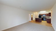 This 2-bedroom, 2-bathroom residence offers a well-designed single-floor layout that maximizes both space and functionality. The open-concept living and kitchen area serves as the heart of the home, with a seamless transition between the carpeted living space and the hard-surface flooring of the kitchen. The kitchen is equipped with rich, dark wood cabinetry, a central island that provides additional prep and dining space, and black appliances including a refrigerator, microwave, and range. Recessed and flush-mount lighting keeps the kitchen bright and functional. The living area benefits from a neutral color palette with clean white walls and a light-toned ceiling, creating an airy and inviting atmosphere. Visible floor vents suggest a reliable climate control system throughout. A hallway leads to the two bedrooms and bathrooms, offering a practical layout that separates the private areas from the main living space. One bathroom is partially visible, hinting at well-appointed finishes consistent with the rest of the unit. Overall, this residence combines a practical, open floor plan with comfortable finishes, making it a functional and welcoming space.