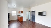 This single-floor unit features a clean, modern design characterized by crisp white walls, smooth ceilings with recessed lighting, and rich dark hardwood-style flooring throughout. The open-concept layout seamlessly connects the living area to the kitchen, creating a sense of spaciousness and ease of movement.

The kitchen is equipped with stainless steel appliances, including a gas range, over-the-range microwave, and a full-size refrigerator. Warm-toned wood cabinetry provides storage above and below, and a compact sink with a granite-style countertop adds functionality. Natural light filters in through multiple windows fitted with horizontal blinds, brightening both the kitchen and living areas.

The living space is generously open, with a wall-mounted flat-screen television serving as a focal point. A wall-mounted air conditioning unit provides climate control. The bathroom is efficiently designed, featuring a glass-enclosed walk-in shower with tile flooring, a toilet, and a window that introduces natural light and ventilation. The unit's overall aesthetic is clean and contemporary, with a neutral palette that offers a versatile, move-in-ready environment.
