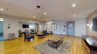 Located at 2751 N 47th St in Philadelphia, PA, this communal lounge space spans a single floor and is thoughtfully designed to accommodate both relaxed gatherings and quiet downtime. The open layout is anchored by light hardwood flooring that runs throughout, creating a warm and cohesive atmosphere across the two distinct areas.

The main lounge area features a generous seating arrangement with contemporary blue armchairs, cushioned ottomans, and wooden coffee tables arranged atop a patterned area rug. A wall-mounted flat-screen television serves as a focal point, complemented by wall-mounted sconce lighting and recessed ceiling lights that provide a bright, welcoming environment. A wall-mounted mini-split air conditioning unit ensures year-round climate comfort.

Framed artwork in coordinating tones lines the light gray walls, adding a refined, curated aesthetic. A secondary, more intimate seating zone offers a quieter setting with additional chairs and side tables, also centered around a wall-mounted television.

An adjacent corridor, accessible through glass sliding doors, features distinctive blue and gold patterned carpet tiles and flush-mount ceiling lighting, connecting this space to the broader building. A hand sanitizer dispenser is conveniently mounted near the entrance. The overall ambiance is clean, modern, and functional, making it an inviting communal retreat.