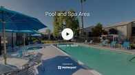Located at 1025 South Gilbert Street in Hemet, California, this outdoor recreational area serves as a central amenity for the surrounding residential community. The centerpiece of the space is a generously sized swimming pool with clear, blue-green water, designed for both lap swimming and leisure. Positioned adjacent to the pool is a circular spa, offering a dedicated relaxation option separate from the main pool. The expansive concrete pool deck is well-appointed with multiple lounge chairs and seating areas, complemented by vibrant blue patio umbrellas that provide shade throughout the day. A wrought iron perimeter fence encloses the pool area, ensuring safety and defined boundaries. Mature landscaping enhances the atmosphere, with towering palm trees and a large weeping willow providing natural shade and a sense of privacy. The surrounding residential buildings, finished in neutral gray tones, frame the space and contribute to a cohesive community feel. Abundant natural light from the Southern California sun fills the space year-round, making it an inviting outdoor retreat. The overall ambiance is relaxed and resort-like, offering residents a welcoming environment for outdoor recreation and leisure.