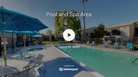 Located at 1025 South Gilbert Street in Hemet, California, this outdoor recreational area serves as a central amenity for the surrounding residential community. The centerpiece of the space is a generously sized swimming pool with clear, blue-green water, designed for both lap swimming and leisure. Positioned adjacent to the pool is a circular spa, offering a dedicated relaxation option separate from the main pool. The expansive concrete pool deck is well-appointed with multiple lounge chairs and seating areas, complemented by vibrant blue patio umbrellas that provide shade throughout the day. A wrought iron perimeter fence encloses the pool area, ensuring safety and defined boundaries. Mature landscaping enhances the atmosphere, with towering palm trees and a large weeping willow providing natural shade and a sense of privacy. The surrounding residential buildings, finished in neutral gray tones, frame the space and contribute to a cohesive community feel. Abundant natural light from the Southern California sun fills the space year-round, making it an inviting outdoor retreat. The overall ambiance is relaxed and resort-like, offering residents a welcoming environment for outdoor recreation and leisure.