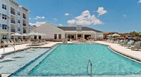 Located at 8920 E College Pointe Dr in Fort Myers, Florida, this single-story community amenity center showcases a thoughtfully designed resort-style outdoor environment. The centerpiece is a generously sized swimming pool with crystal-clear turquoise water, surrounded by an expansive paved pool deck finished in light-toned pavers that reflect the Florida sunshine. Rows of chaise lounges and shade umbrellas in soft sage green tones create a relaxed, resort atmosphere perfect for enjoying the warm Southwest Florida climate.

The clubhouse building itself features a striking modern farmhouse aesthetic, combining crisp white board-and-batten siding with a stone veneer base, a standing-seam metal roof, and a prominent stone chimney. Large windows allow natural light to flow through the interior while offering views of the pool area. Outdoor seating areas with plush lounge furniture and accent pillows in neutral and aqua tones are positioned along the building's exterior.

The pool deck extends to a waterfront promenade lined with lounge chairs and umbrella shading, offering serene views of an adjacent pond framed by lush landscaping. Restroom facilities and a water fountain are conveniently accessible from the deck. A covered porch area with an outdoor TV and stylish seating provides an additional space for relaxation and entertainment, completing this comprehensive outdoor amenity offering.