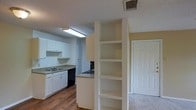 This single-floor space features an open-concept design that connects the kitchen and living areas seamlessly. The kitchen is outfitted with white cabinetry providing ample storage, a black dishwasher, a gas range, and a sink set into a light-toned countertop. A skylight-style fluorescent fixture illuminates the kitchen workspace, enhancing the bright, clean aesthetic created by the white cabinetry. A half-wall partition separates the kitchen from the adjoining area and incorporates a tall built-in shelving unit with multiple open shelves, offering practical display or storage space. The flooring transitions from warm hardwood-style planks in the kitchen and main area to a lighter surface near the doorway, visible to the right. Walls are painted in a neutral yellow-beige tone, complementing the white trim and doors throughout. The textured ceiling adds a classic residential character to the space. A standard interior door on the right side of the image leads to an additional room, suggesting a functional layout suited for everyday living.