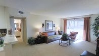 This single-floor residence offers a welcoming and well-appointed living space designed with comfort and functionality in mind. The living room serves as the heart of the home, showcasing a warm, neutral color palette with beige carpeted flooring and smooth, light-toned walls that create a calm and inviting atmosphere. A large window dressed with rust-colored curtains allows generous natural light to fill the room, offering views of the outdoors and enhancing the sense of openness. The space is thoughtfully furnished with a plush sofa accented by colorful throw pillows, a leather armchair, and a pair of round nesting coffee tables, all arranged to encourage relaxed conversation and everyday living. Wall sconces and table lamps provide warm ambient lighting, complementing the framed artwork displayed on the main wall. Lush indoor plants add a refreshing touch of greenery throughout the space. A hallway visible from the living area suggests additional rooms and functional spaces beyond, indicating a practical and well-organized floor plan. The textured ceiling and clean architectural lines contribute to the overall understated yet polished aesthetic of this comfortable single-story home.